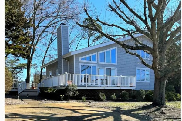 Cape Cod-style house with gray shingles, white trim, two dormers, front porch, and manicured lawn surrounded by trees.